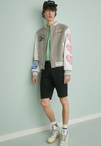 Veste varsity grise et blanche avec des écussons, chemise verte, shorts noirs, baskets blanches et casquette noire. Tissu en coton, poignets et col côtelés.