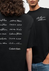Deux personnes portant des t-shirts noirs Calvin Klein, l'une affichant plusieurs impressions de logos blancs au dos, l'autre un logo blanc à l'avant.