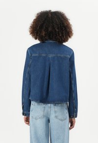 Temno modra džins jakna s sproščenim krojem, ki vključuje zadaj pregib, zapenjanje z gumbi na rokavih in ravno spodnjo rob. Ujemajoča se z svetlo modrimi jeans hlačami.