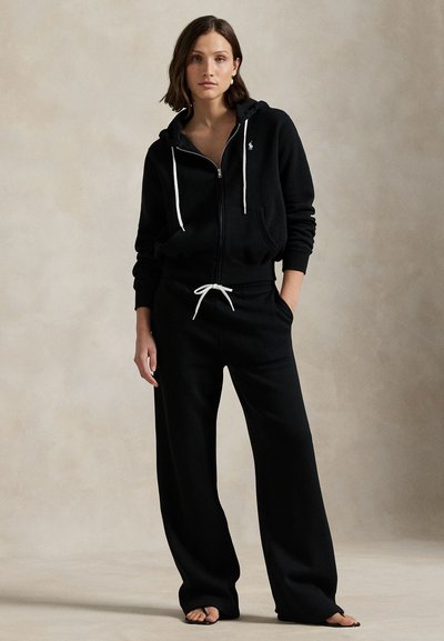 Žena stojící s rukama v kapsách, oblečená v černé mikině na zip a širokých kalhotách s bílými šňůrkami, bosá, na neutrálním pozadí.