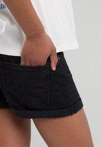 Black denim shorts with rolled hem, featuring a back pocket and a textured surface. The model has their hand in the pocket.