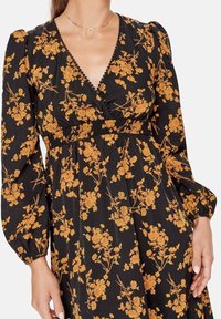 Kvinna klädd i en svart klänning med orange blommönster, V-ringning, puffärmar och elastisk midja, visar överkropp och händer.