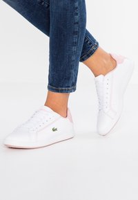 Baskets en cuir blanc avec accents roses, design à lacets, profil bas, semelle en caoutchouc texturée et logo brodé sur le côté.