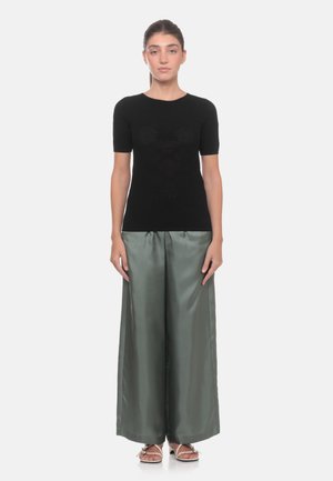 Donna in piedi di fronte che indossa una maglietta nera aderente a maniche corte, pantaloni larghi grigi lucidi e sandali aperti sulle dita.