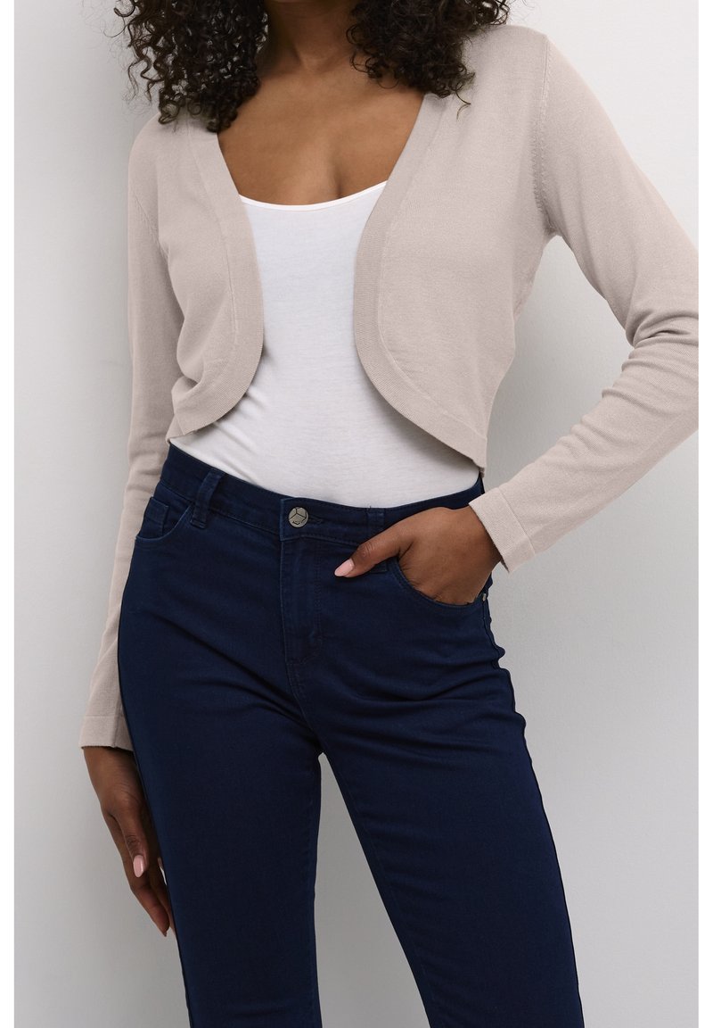 Femme portant un cardigan court beige sur un haut blanc avec un jean taille haute bleu foncé, une main dans la poche, sur fond blanc.