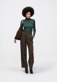 Frau mit Afro, die einen dunkelgrünen Rollkragenpullover, hochgeschnittene braune weit geschnittene Hose und schwarze Stiefel trägt, während sie eine große braune Schultertasche trägt. Sie steht vor einem weißen Hintergrund.