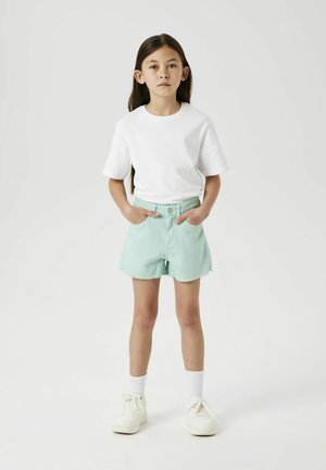T-shirt blanc à manches courtes associé à un short en denim vert menthe avec un ourlet effiloché. Des baskets blanches et des chaussettes mi-mollet viennent compléter la tenue.