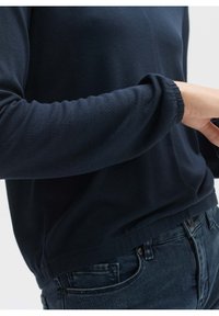 Námořnická modrá dlouhá rukáv pulovr z měkké, texturované látky; má nařasené manžety a volný střih v pase, spojený s tmavými džínami.