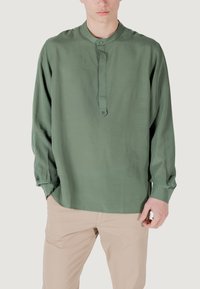 Camicia verde a maniche lunghe con colletto alla coreana, patta frontale con bottoni, texture morbida e vestibilità rilassata, abbinata a pantaloni beige chiaro.