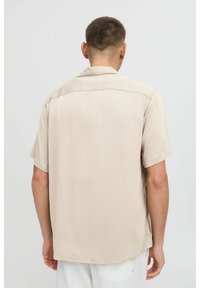 Korte mouwen, beige shirt gemaakt van een lichtgewicht stof. Heeft een klassieke kraag en een gladde textuur. Achteraan is een eenvoudig ontwerp te zien.