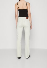 Jeans blancos acampanados con una cintura alta, diseñados en denim. La vista trasera muestra dos bolsillos traseros y un parche de logotipo de cuero marrón en la cintura.