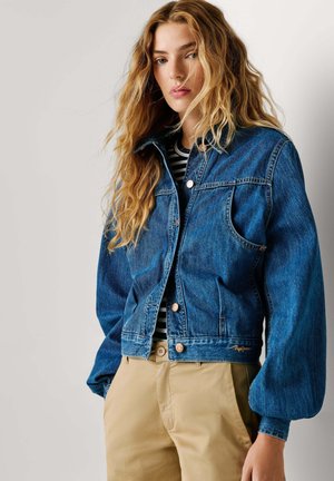 Junge Frau mit langen, welligen Haaren, die eine blaue Jeansjacke über einem schwarz-weiß gestreiften Hemd und beigen Hosen trägt, vor einem schlichten Hintergrund stehend.