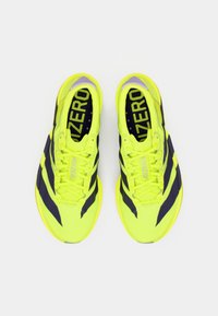 Αθλητικά παπούτσια με έντονο κίτρινο επάνω μέρος, σκούρες μοβ ρίγες και υφή υφάσματος. Διαθέτουν κορδόνια και μαλακές εσωτερικές σόλες.