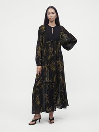 Femme aux longs cheveux noirs portant une robe maxi noire à manches longues à motif floral et des sandales à talons noires, debout devant un fond uni.