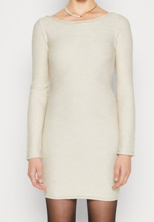 Cremefarbenes, langärmeliges Strickkleid mit Bootsausschnitt, kombiniert mit transparenten schwarzen Strumpfhosen und einer unpassenden Halskette aus Perlen und Goldkette.