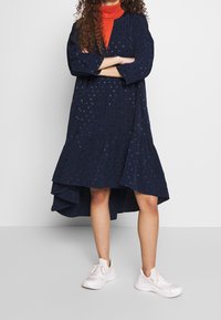 Mörkblå klänning med utsvängd nederkant, prydd med subtila prickar och trekvartsärmar, kombinerad med vita sneakers.