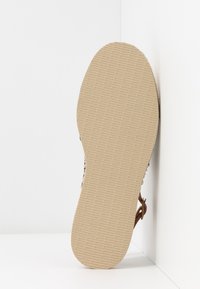 Semelle d'une chaussure plate beige avec un motif de grip texturé, positionnée à la verticale contre un mur d'angle blanc.