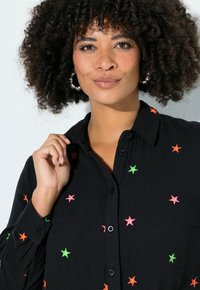 Vrouw met krullend haar, zilveren oorringen in de vorm van ringen en een zwart overhemd met knopen, geborduurd met kleurrijke sterren, waarbij ze met één hand de kraag vasthoudt.