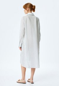 Abito camicia bianco lungo con strisce verticali blu, colletto e maniche lunghe. Tessuto leggero con fessure laterali e vestibilità rilassata.