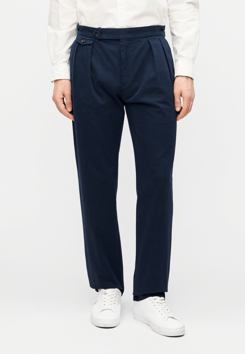 Pantalon en coton bleu marine à coupe ajustée, avec des plis devant, une patte latérale avec un bouton, porté avec des baskets blanches.