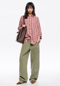 Camisa de cuadros roja y negra de ajuste holgado, combinada con pantalones anchos de color oliva y zapatos puntiagudos marrones, sosteniendo una bolsa marrón oscuro.