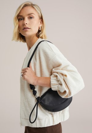 Zwarte leren handtas met een halvemaanvorm en een geknoopte handgreep. Textuur crèmekleurige trui met een ontspannen pasvorm, gedragen over donkergroene broek.