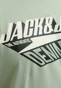 Tricou din bumbac de culoare verde deschis, cu text grafic alb-negru. Designul include tipografie îndrăzneață și un accent geometric.