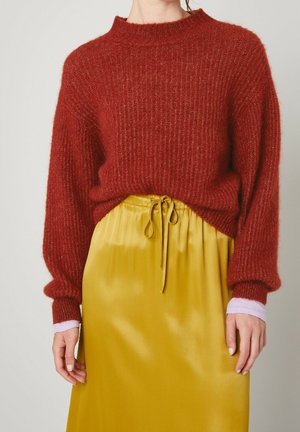 Femme portant un pull rouge texturé à manches bouffantes sur une chemise violette claire, associé à une jupe jaune moutarde brillante avec cordon de serrage.