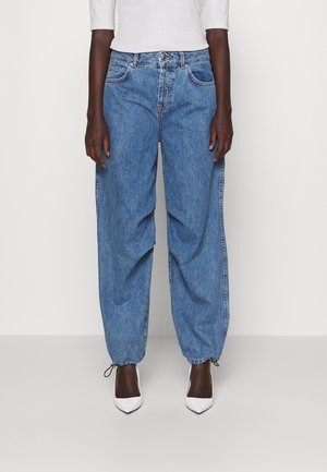 Persoon draagt losse blauwe denim jogger jeans met verstelbare enkelbandjes en witte pumps met een puntige neus, gecombineerd met een lichtgrijze top.