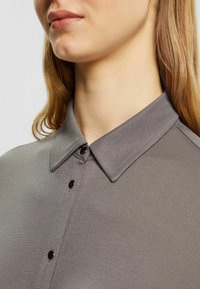 Chemise grise à boutons avec un col classique, présentant des boutons noirs et un tissu doux et texturé. Vue en gros plan des détails du col et des boutons.