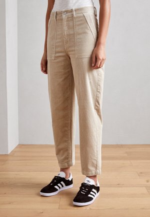 Beige manchesterbukser med vide ben, høj talje og store forlommer kombineret med sorte og hvide sneakers. Trægulv synligt.