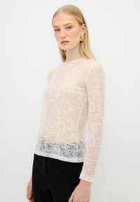 Top in pizzo trasparente con intricate fantasie floreali, scollatura rotonda e maniche lunghe; il colore bianco sfuma in nero all'orlo.