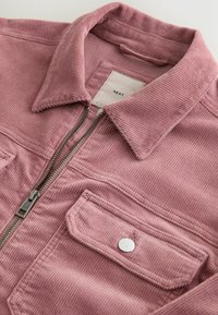 Veste en velours côtelé rose poussiéreux avec col, fermeture éclair et poche à rabat avec bouton blanc sur fond blanc.