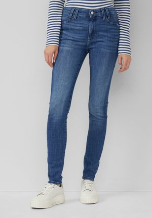 Jean skinny bleu en denim avec une texture lisse, cinq poches et une taille mi-haute, associés à des baskets blanches.
