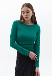 Top aderente a maniche lunghe di colore verde vibrante, realizzato in tessuto morbido. Presenta un collo a barca e una leggera arricciatura sui lati.