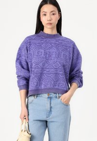 Lila kortärmad sweatshirt med ett abstrakt mönster, rund hals, och överdimensionerade ärmar, kombinerat med ljusblå jeans och bärande på en väska.