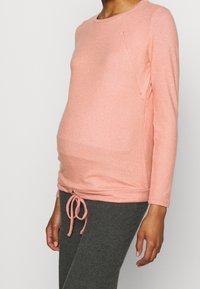 Růžový těhotenský top s dlouhým rukávem a stahovací šňůrkou v pase, s texturovaným povrchem a detaily bočních kapes. Kombinováno se šedými legínami.