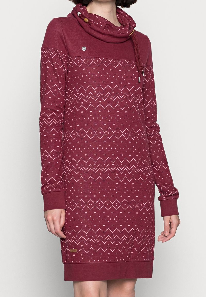 Femme portant une robe à manches longues bordeaux avec des motifs géométriques roses et un col haut et ample avec des cordons.