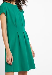Grön kortärmad klänning med rund halsringning, texturerad tyg och paneldetaljer. Klänningslängden vidgar sig, vilket skapar en något A-linjeform.