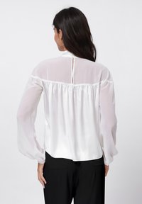 White satin blouse with a high neckline, long sheer sleeves, and a keyhole opening at the back. Smooth texture and flowing design.
