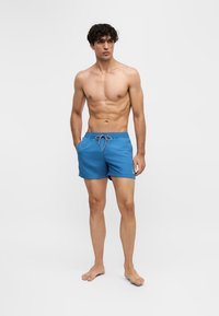 Jeune homme debout pieds nus portant un short de bain bleu avec les mains dans les poches sur un fond clair uni.