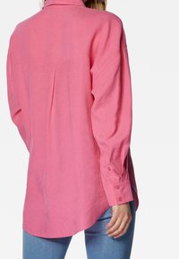 Camicia a maniche lunghe in lino rosa con yoke posteriore, polsini con bottoni e un orlo curvo. Il tessuto sembra leggero e traspirante.
