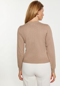 Beige, gerippter Strickpullover mit langen Ärmeln und Rundhalsausschnitt, mit einer engen Saum und weicher Textur, getragen mit weißen Hosen.
