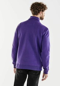 Purple sweatshirt with a high collar, ribbed cuffs, and a textured finish, paired with dark jeans. Clean lines and casual design.