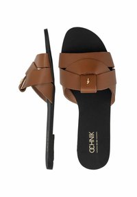 Brązowe klapki z sztucznej skóry z szerokim paskiem i małym złotym emblematem, spoczywające na czarnej, textured wkładce. Płaska podeszwa.