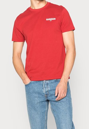 Mężczyzna ubrany w czerwoną koszulkę z krótkim rękawem z małym białym logo na piersi i jasnoniebieskie dżinsy, stojący z jedną ręką w kieszeni.