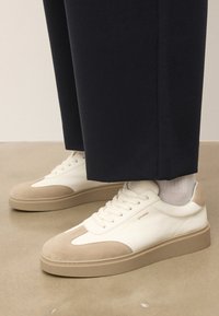 Baskets basses blanches et beiges avec un design à lacets, portées avec des chaussettes blanches et un pantalon noir, se tenant sur un sol beige.