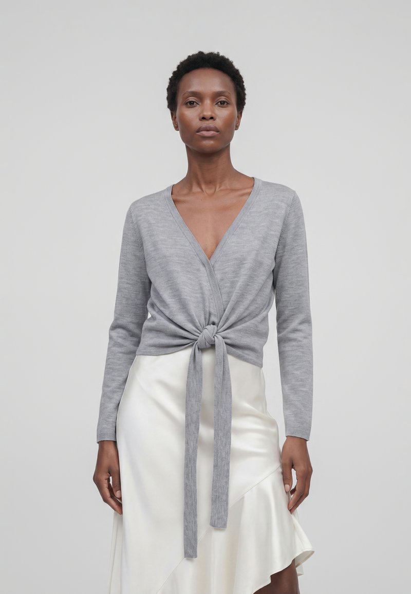 Femme portant un pull cache-cœur gris clair à manches longues noué devant et une jupe asymétrique en satin blanc, debout devant un fond uni.