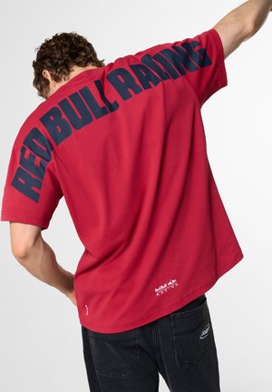 Mand iført en rød T-shirt med stor sort "REDBULL RACING" tekst på ryggen og lille Red Bull Racing-logo nær kanten.