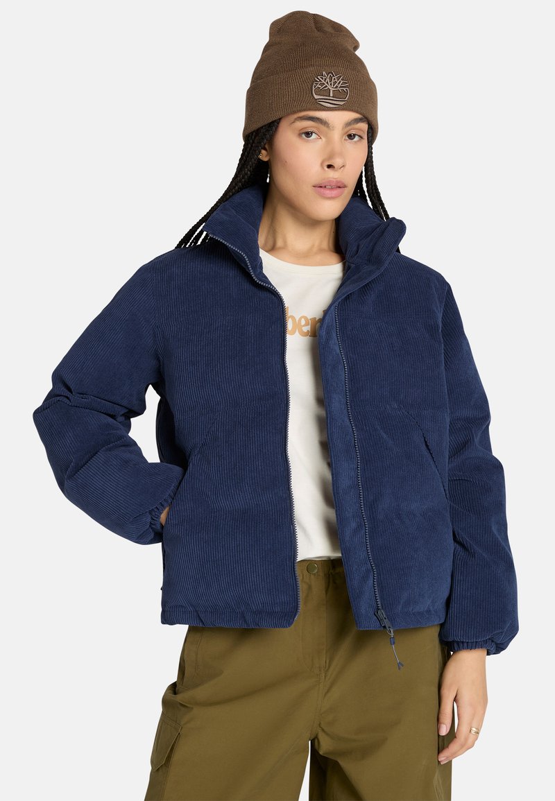 Marineblå fløjlsjakke med høj krave, frontlynlås og elastiske manchetter. Parret med en beige skjorte og olivengrønne bukser. Brun hue med logo.